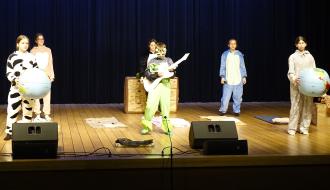 Grupa dzieci i młodzieży ze Szkoły Podstawowej w Idzikowicach występuje na scenie RCK w kostiumach typu kigurumi (jednoczęściowe piżamy zwierzęce). Po lewej i prawej stronie stoją osoby trzymające duże, nadmuchiwane modele kuli ziemskiej. W centralnej części młody artysta gra na białej gitarze elektrycznej. 