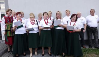 Na zewnątrz, przed szarą ścianą budynku, stoi zespół ludowy z KGW Idzikowice składający się głównie ze starszych kobiet. Kobiety w większości ubrane są w białe bluzki z logo, ciemnozielone spódnice i czerwone korale, trzymając w rękach śpiewniki. Jedna osoba nosi tradycyjny, kolorowy strój ludowy.