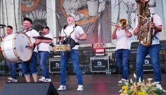 Zespół muzyczny GiS w białych koszulkach i dżinsach grający na instrumentach dętych i perkusyjnych na scenie.