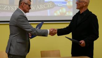 Burmistrz Drzewicy i Prezes Fundacji Edukacji Przedszkolnej w garniturach podają sobie dłonie, trzymając w rękach pamiątkowe teczki z dokumentami. W tle widoczny jest ekran projekcyjny z prezentacją.
