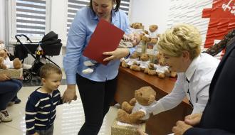 Pani Dyrektor CUS wręcza pluszowego misia w pudełku małemu chłopcu stojącemu obok mamy z niemowlęciem na rękach. 