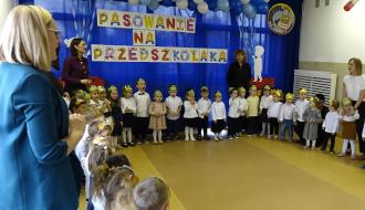 Widok ogólny na salę przedszkolną, gdzie dzieci w odświętnych strojach stoją w półkolu z wychowawczyniami. W tle napis „PASOWANIE NA PRZEDSZKOLAKA” oraz dekoracja z balonów.