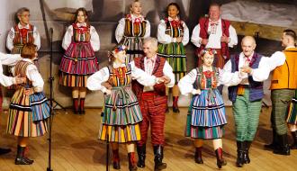 Pary taneczne ZPiT „Drzewiczanie” w barwnych, tradycyjnych strojach ludowych wykonują taniec na drewnianej scenie. W tle stoi grupa śpiewacza oraz muzycy w strojach regionalnych, obserwujący występ.