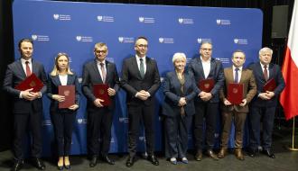 Dziewięć osób ustawionych w rzędzie na tle niebieskiej ścianki z logo Ministerstwa Infrastruktury. W tym gronie m.in. Minister Infrastruktury, Wojewoda Łódzki i Burmistrz Drzewicy. Większość trzyma czerwone teczki