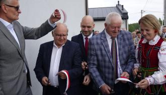 Kolejne ujęcie z momentu przecięcia wstęgi na zewnątrz. Burmistrz Drzewicy - po lewej stronie - podnosi odcięty kawałek biało-czerwonej taśmy do góry.