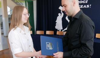 Zastępca Burmistrza Drzewicy w czarnej koszuli wręcza niebieski dyplom i uściskiem dłoni gratuluje uczennicy w okularach.