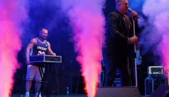 Wokalista zespołu D-Bomb w garniturze i DJ przy konsolecie na oświetlonej na różowo scenie z efektami dymnymi.