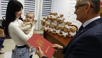 Burmistrz Drzewicy w garniturze wręcza czerwony folder z herbem Drzewicy matce trzymającej na rękach małe dziecko.