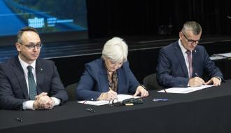 Siedząc umowę podpisują Wojewoda Łódzki Dorota Ryl i Burmistrz Drzewicy Janusz Reszelewski. Obok po lewej stronie siedzi Minister Infrastruktury Dariusz Klimczak. W tle część ekranu oraz ciemna przestrzeń.