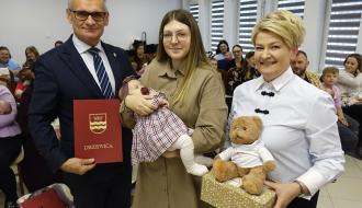 Pozowane zdjęcie do pamiątki: młoda matka z niemowlęciem stoi pomiędzy Burmistrzem Drzewicy w garniturze z czerwoną teczką a Panią Dyrektor CUS trzymającą maskotkę misia na pudełku. 