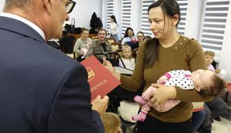 Burmistrz Drzewicy w garniturze wręcza czerwoną teczkę z herbem Drzewicy matce trzymającej na rękach śpiące niemowlę. Obok kobiety stoi małe dziecko.