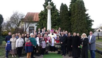 Pamiątkowe zdjęcie grupowe uczestników uroczystości na tle odrestaurowanej Kolumny Toskańskiej i zielonych drzew. W środku stoi Arcybiskup Wacław Depo, w otoczeniu licznych mieszkańców, osób w strojach ludowych, dzieci oraz zaproszonych gości.