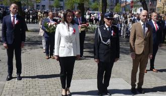 W parku na Placu Wolności ceremonia składania wieńca. Na pierwszym planie Przewodnicząca Rady Miejskiej w Drzewicy, Burmistrz w galowym mundurze OSP i Zastępca Burmistrza Drzewicy. Wszyscy z biało-czerwonymi kotylionami. W tle pozostali uczestnicy wydarzenia, część trzyma wiązanki kwiatów, dalej zabudowa miejska i drzewa.