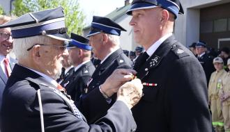 Druh Tadeusz Cisowski przypina odznakę strażacką druhowi Jackowi Dziurzyńskiemu. W tle inni strażacy i po prawej budynek OSP Drzewica.