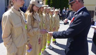 Burmistrz Janusz Reszelewski w galowym mundurze OSP przekazuje odznakę jednej z członkiń MDP Domaszno. Obok druhny po lewej jedna, a po prawej pięć dziewcząt również ubranych w jasne mundury OSP. 