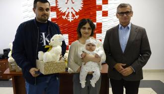 Od lewej stoją: tata trzymający pluszowego misia i wyprawkę, mama trzymająca bobasa oraz Burmistrz Drzewicy Janusz Reszelewski.