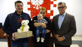 Od lewej stoją: tata trzymający pluszowego misia i wyprawkę, mama trzymająca bobasa oraz Burmistrz Drzewicy Janusz Reszelewski.
