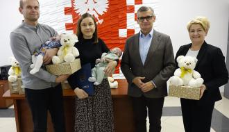 Od lewej stoją:  tata trzymający pluszowego misia, wyprawkę i bobasa, mama trzymająca bobasa oraz Burmistrz Drzewicy Janusz Reszelewski i Dyrektor MGOPS Marta Woźniak