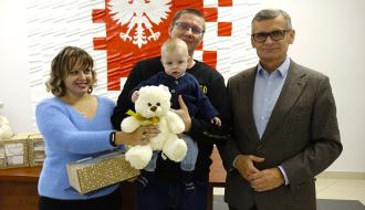 Od lewej stoją: mama trzymająca wyprawkę i pluszowego misia, tata trzymający bobasa i Burmistrz Drzewicy Janusz Reszelewski.