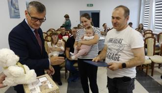 Burmistrz Drzewicy Janusz Reszelewski przekazuje prezent, dyplom i maskotkę tacie małego dziecka. Obok mama trzyma bobasa na rękach.