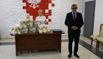 Sala USC Urzędu Miejskiego w Drzewicy. Na tle biało czerwonej ściany z godłem Polski Burmistrz Drzewicy Janusz Reszelewski oraz Dyrektor MGOPS w Drzewicy Marta Wożniak. Na stole przed Panią Dyrektor ustawione wyprawki dla najmłodszych mieszkańców Gminy Drzewica. 