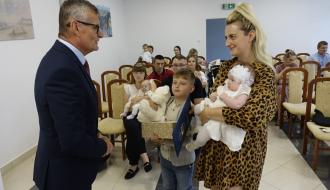 Burmistrz Drzewicy Janusz Reszelewski przekazuje prezent, dyplom i maskotkę mamie małego dziecka. Kobieta trzyma bobasa na rękach. Obok starszy brat malucha. 