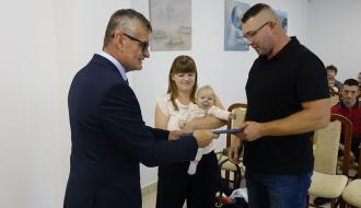 Burmistrz Drzewicy Janusz Reszelewski przekazuje dyplom tacie  małego dziecka. Z tyłu mama trzymająca na rękach dziecko. 