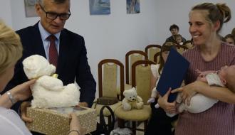 Burmistrz Drzewicy Janusz Reszelewski przekazuje prezent, dyplom i maskotkę mamie małego dziecka. Kobieta trzyma bobasa na rękach.