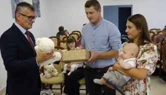 Burmistrz Drzewicy Janusz Reszelewski przekazuje prezent, dyplom i maskotkę tacie małego dziecka. Obok mama trzyma bobasa na rękach.