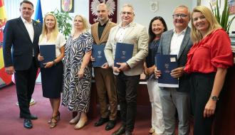 Grupa ośmiu osób w gabinecie pozuje do wspólnego zdjęcia. Cztery osoby prezentują niebieskie teczki z podpisanymi dokumentami. W środku Zastępca Burmistrza Drzewicy Dominik Niemirski.