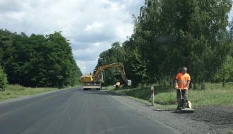 Prace związane z wykonaniem utwardzenia pobocza oraz odtworzenia rowów. Na pierwszym planie zdjęcia pracownik obsługujący ugniatarkę w tle koparka podczas prac. 