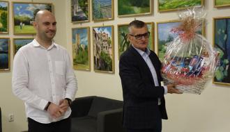 Na zdjęciu Burmistrz Drzewicy Janusz Reszelewski i Zastępca Burmistrza Dominik Niemirski z życzeniami z okazji Święta Pracownika Kultury w Regionalnym Centrum Kultury w Drzewicy. Burmistrz dzierży kosz słodyczy, obok niego Zastępca. W tle na ścianie obrazy na których widać drzewickie pejzaże