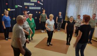 Grupa seniorów na zajęciach fitness w SP w Idzikowicach. Na pierwszym planie instruktor zajęć Alina Szymańska, na planie dalszym uczestnicy zajęć patrzący na instruktora wykonują ćwiczenia. 