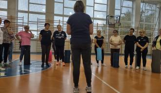 Grupa seniorów na zajęciach fitness w hali sportowej SP w Radzicach Dużych. Na pierwszym planie instruktor zajęć Maria Smolarska, na planie dalszym uczestnicy zajęć patrzący na instruktora