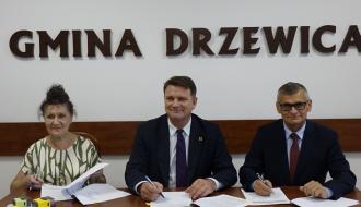 1.	W sali konferencyjnej Urzędu Miejskiego w Drzewicy, przy stole, na tle ściany z napisem Gmina Drzewica, herbem Drzewicy oraz godłem Rzeczpospolitej Polskiej siedzą: (od lewej) Skarbnik Gminy Drzewica Irena Jóźwicka, Wice Marszałek Województwa Łódzkiego Artur Bagieński oraz Burmistrz Drzewicy Janusz Reszelewski. 