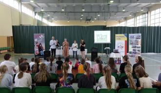 Na zdjęciu uczniowie trzecich klas szkół podstawowych z terenu Gminy Drzewica znajdują się w hali sportowej SP w Drzewicy przy ul. Staszica. Hala przedzielona jest kotarami, na parkiecie rozstawione rollupy promocyjne szkoły oraz firmy Orto Med. Sport w Łodzi. Na stoliku przyktytym białym obrusem  ustawione rollery. Przed uczniami stoją Zastępca Burmistrza Dominik Niemirski, Dyrektor SP w Drzewicy Agnieszka Bogatek oraz przedstawicielki firmy Ortomed w Łodzi