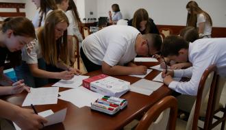 Grupa młodzieży pochylona nad stołem wypełnia kartki i notuje. Na stole leżą markery i kartki