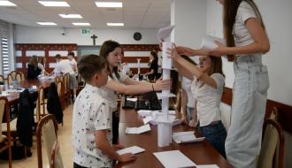 Grupa młodzieży pracuje przy stole, wspólnie budując konstrukcję z papieru. W tle inni uczestnicy wykonują podobne zadania w sali konferencyjnej