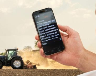 Na pierwszym planie dłoń a w niej telefon z tekstem wiadomości, w tle traktor na polu