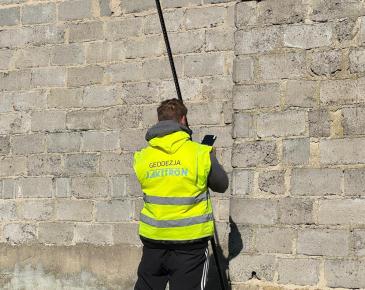 Mężczyzna w kamizelce odblaskowej wykonuje pomiar przy ścianie budynku za pomocą sprzętu geodezyjnego.