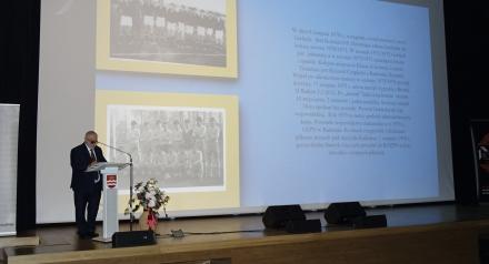 Po lewej stronie stoi starszy mężczyzna w garniturze, który przemawia przy mównicy z mikrofonem. Przed mównicą znajduje się bukiet kwiatów. Za nim na dużym ekranie wyświetlana jest prezentacja. 