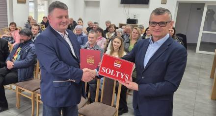 Burmistrz wręcza teczkę ora tabliczkę z napisem sołtys Sołtysowi Augustowa w tle mieszkańcy