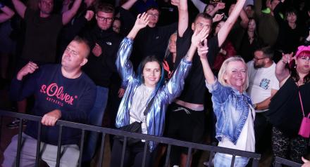 Grupa bawiących się ludzi przy barierce podczas imprezy plenerowej, unosząca ręce w geście radości. 