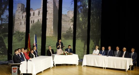 grupa osób siedzących w sali przy stołach z białymi obrusami oraz wielką grafiką w tle przedstawiającą ruiny zamku drzewieckiego, obchodzi ceremonię rocznicy nadania Drzewicy praw miejskich 
