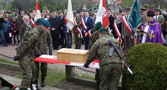 żołnierze składają szczątki poległych przedstawicieli Wojska Polskiego. W tle żołnierze trzymający flagi z godłem polski, ksiądz wraz z grupą ludzi uroczyście ubranych.