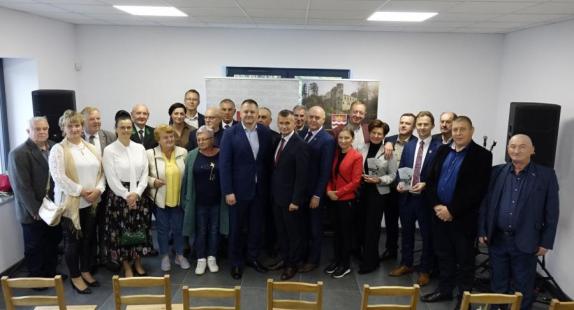 dwudziestokilkuosobowa grupa osób ustawiona po jednej stronie sali podczas uroczystości otwarcia wyremontowanego „Domku Wędkarza” w Drzewicy