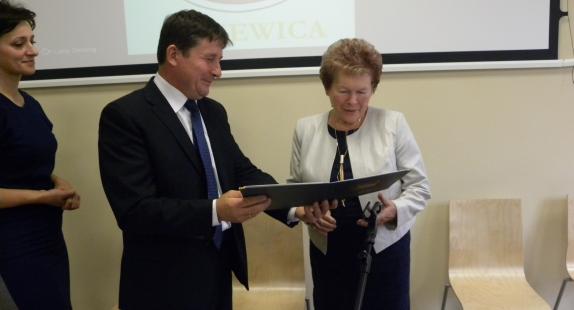 Na zdjęciu starsza kobieta trzyma dokument i przygląda się mu z uwagą. Obok niej znajduje się mężczyzna oraz kobieta, którzy wręczają jej ten dokument.. W tle widoczny jest ekran projekcyjny z herbem Gminy Drzewica 