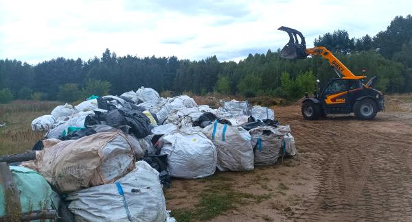 Ułożone na ziemi spakowane w baloty folie i inne odpady rolnicze. Obok po prawej ciągnik-ładowarka na piaszczystym podłożu. W tle las.