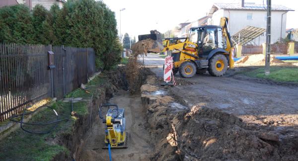 Przebudowa sieci wodociągowej na Os. Słonecznym w Drzewicy. Na zdjęciu wykop, w środku którego znajduje się zagęszczarka. W tle koparko-ładowarka zasypuje wykop drobnym piaskiem. Wokoło zabudowania przy ulicy. 