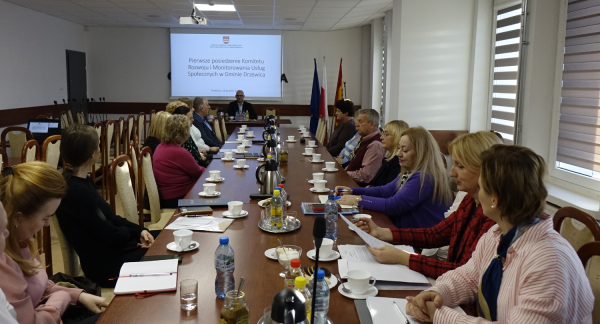 Spotkanie komitetu w Sali konferencyjnej Urzędu Miejskiego w Drzewicy. Na pierwszym planie przy stole głównym członkowie komitetu. W tle przy stole prezydialnym Zastępca Burmistrza Drzewicy Dominik Niemirski. Na ścianie za jego plecami na ekranie prezentacja multimedialna. 